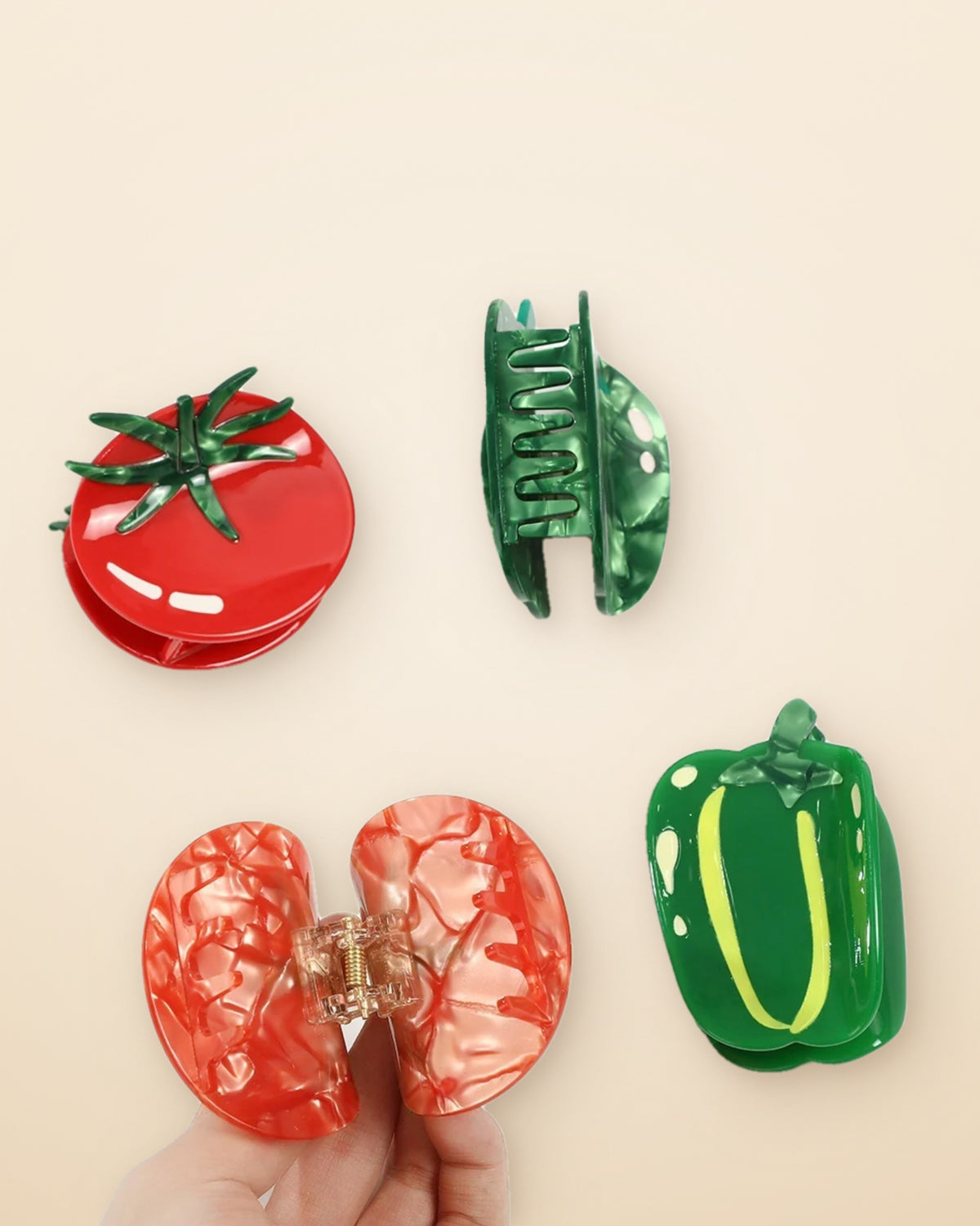 Hand opening a marbled tomato claw clip beside tomato and green pepper clips on a sand background