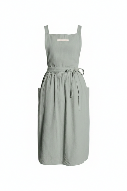 A sage green linen apron with square shoulder straps, a tie waist belt, and two front pockets, shown against a plain white background.