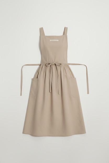 A light beige linen apron with a tie waist and front pockets, displayed flat against a light gray background.
