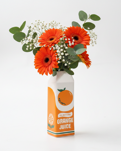 A floral arrangement of bright orange gerbera daisies, green eucalyptus leaves, and small white baby's breath stems, styled in the shape of an orange juice carton on a clean white background.