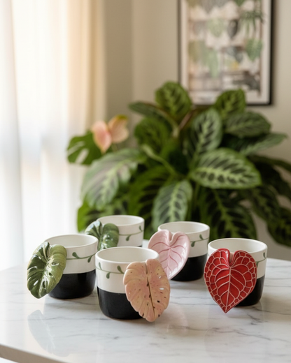 Six ceramic plant pot holders shaped like different tropical leaves, arranged on a white marble table in front of a green houseplant. The pots are black and white with the leaf design, and the leaves are in shades of green, pink, and red.