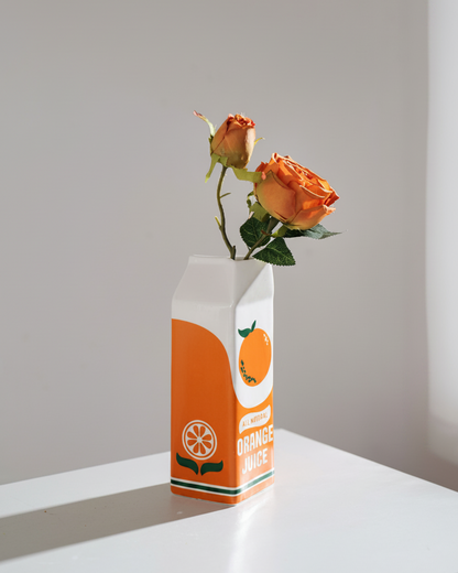 An orange and white juice carton vase holding two orange roses on a white table against a neutral gray background.