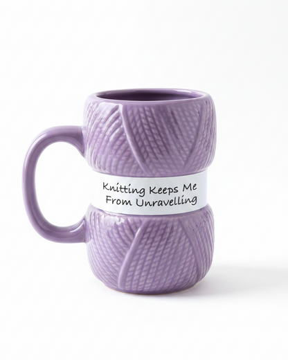 A purple ceramic mug designed to look like a ball of yarn, featuring a white paper band wrapped around the middle with the text 'Knitting Keeps Me From Unravelling' in black script.