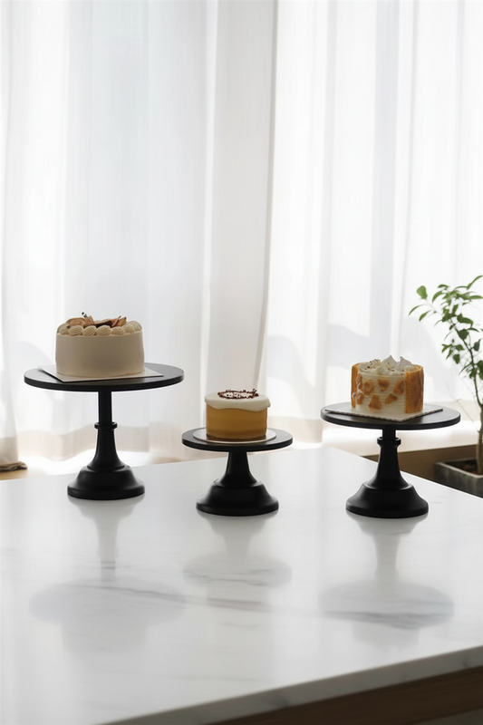 Three black round cake stands with pedestal bases are arranged on a white marble countertop in a bright kitchen setting, holding three decorated cakes in front of sheer white curtains.