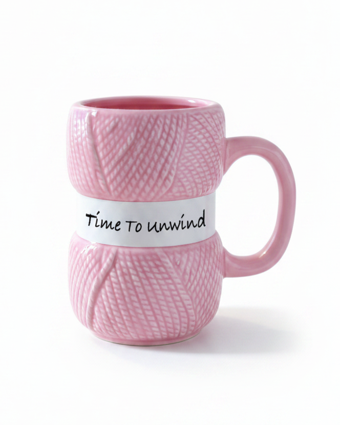 A pink ceramic mug designed to look like a spool of yarn, featuring a white band around the middle with the handwritten text 'Time to unwind'.