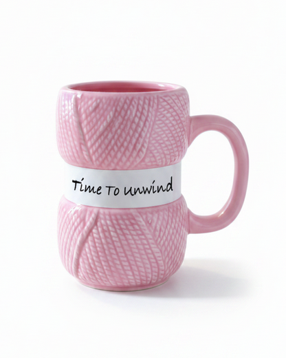A pink ceramic mug designed to look like a spool of yarn, featuring a white band around the middle with the handwritten text 'Time to unwind'.