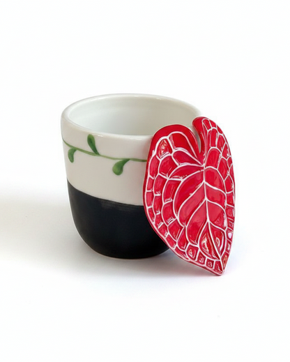A small white ceramic cup with a black bottom half and green leaf details, featuring a red heart-shaped ceramic lid with white leaf veins leaning against it on a white background.